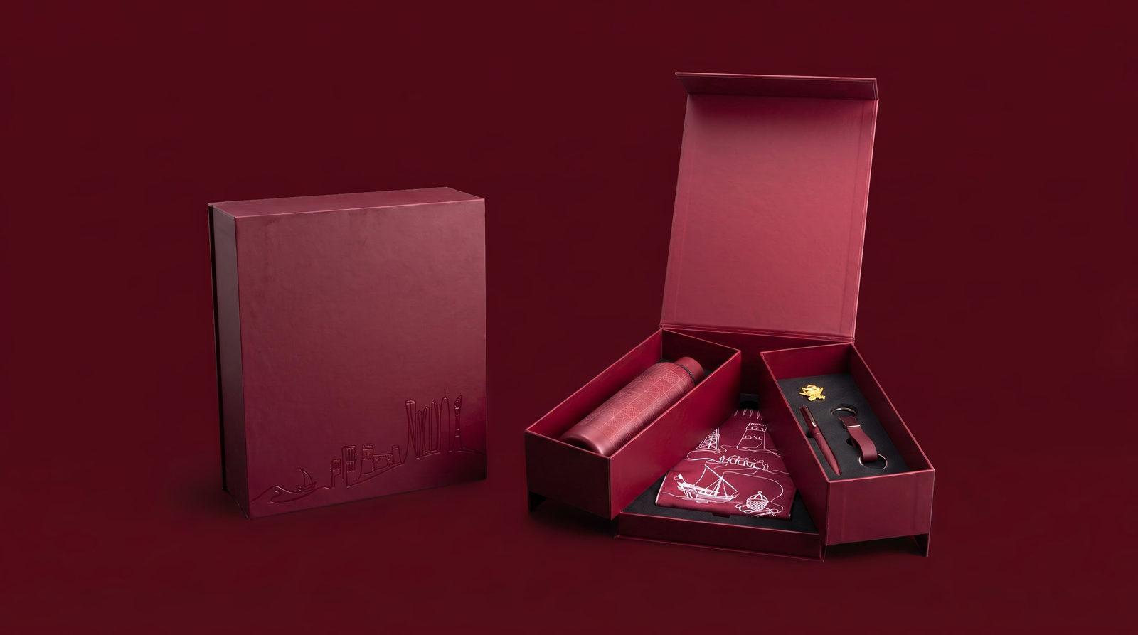 The Fakhr Al-Muwazzaf presentation box open on deep maroon, showing the bottle, scarf, pen, keychain and badge.
