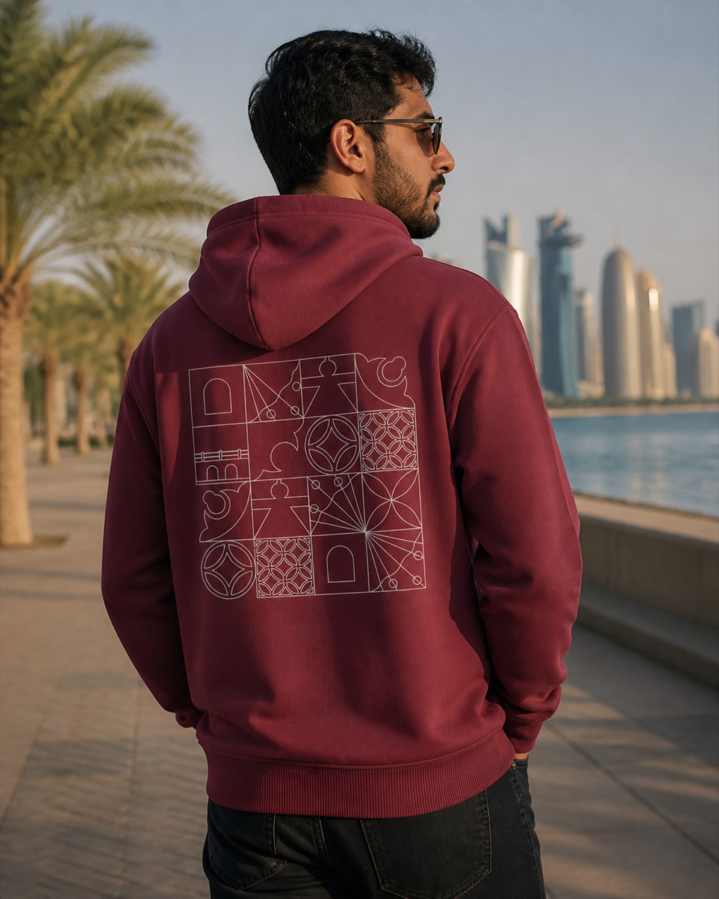 A man on the Doha Corniche at golden hour wearing the Izz Qatar hoodie, its heritage tile pattern across the back.