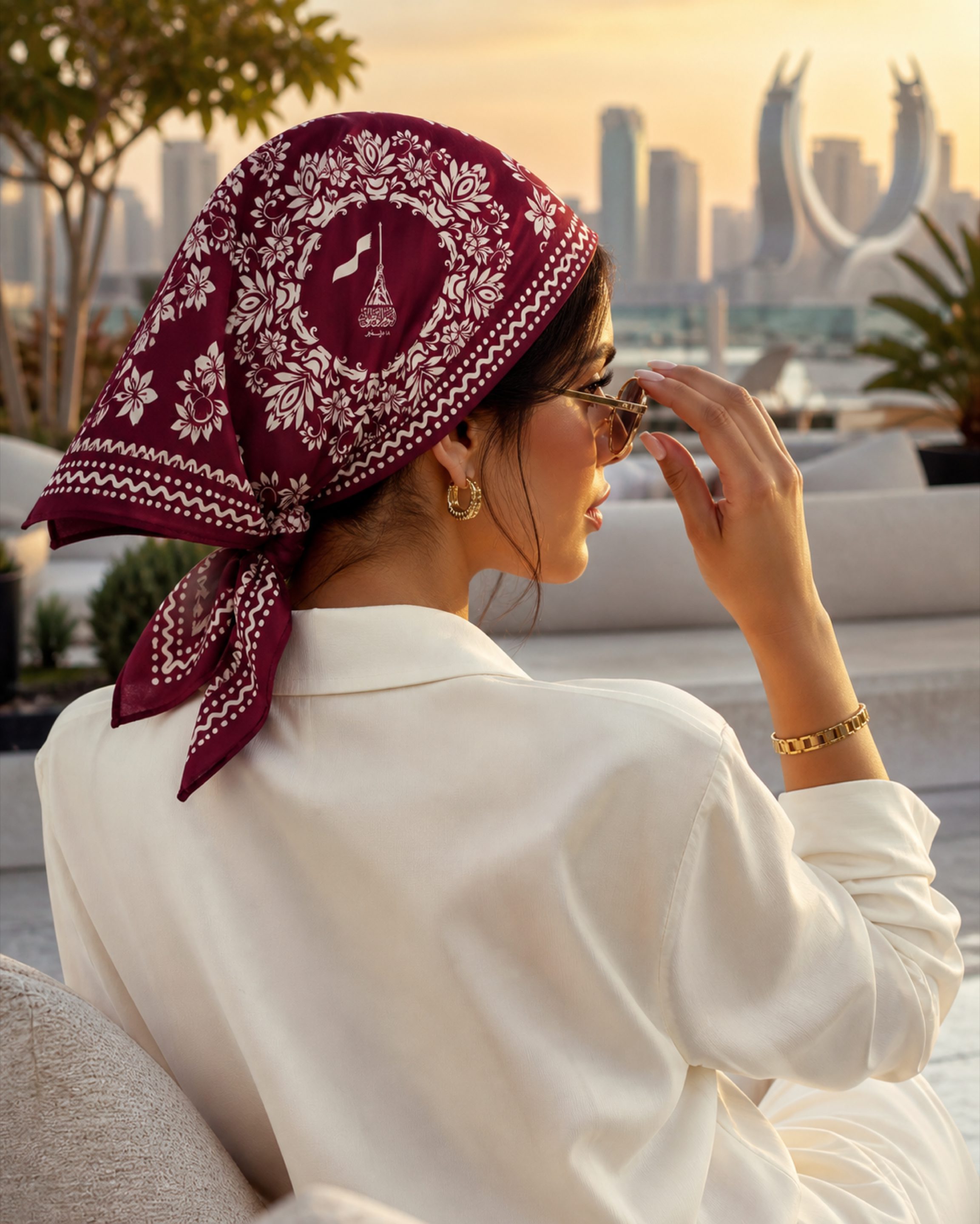 A woman on a Doha rooftop at sunset wearing the Izz Qatar floral bandana, the West Bay skyline behind her.