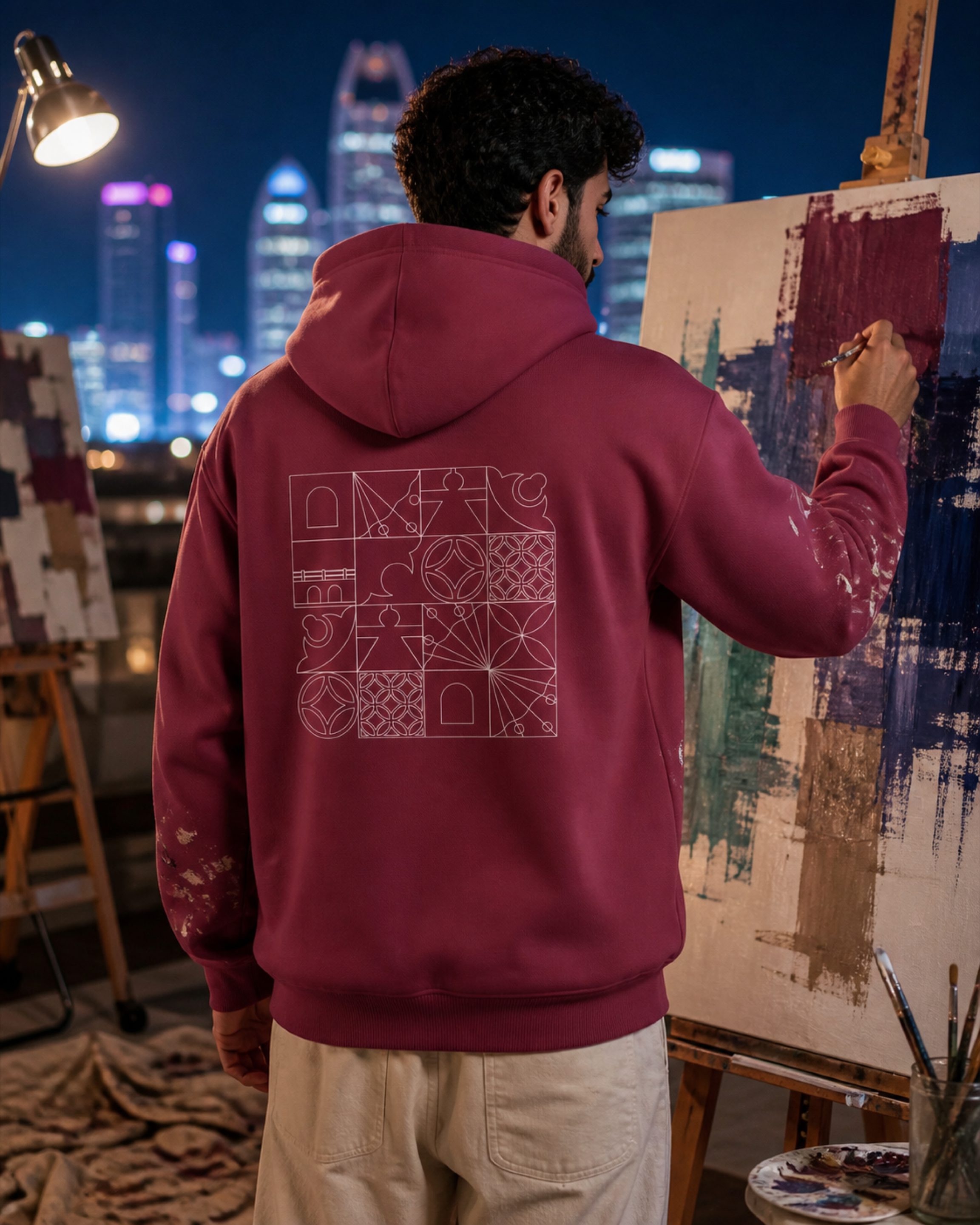 A painter at an easel in a Doha studio at dusk, the Izz Qatar hoodie pattern across his back, the lit skyline through the window.